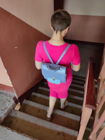 The image shows a person walking down a set of concrete stairs. The person is viewed from behind and is wearing a bright pink, short-sleeved, ribbed dress and a light blue backpack. The backpack has a silver clasp and two blue straps. The person is also wearing light-colored sandals.
The staircase appears to be indoors, likely in an apartment building, with a brown, textured wall on the left and a reddish-brown wooden handrail on the right. The steps are worn and appear to be made of concrete. The lighting is somewhat dim, suggesting it could be daytime, possibly indoors or in a shaded area. There is no visible text in the image. The overall impression is of someone going about their daily routine in a utilitarian setting. The location context is Fanipal, Belarus.