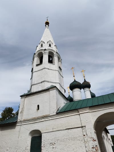 The image shows the exterior of a white brick church with a tall, conical bell tower topped with a golden dome and cross. The bell tower has arched openings and decorative detailing. To the right of the bell tower, several smaller domes with green roofs and golden crosses are visible, characteristic of Russian Orthodox architecture. The church is made of weathered white brick. The sky above is overcast with gray clouds. The scene is an outdoor shot, looking up at the architecture. The location of Шебунино, Russia is mentioned, but the image itself does not contain specific visual cues to confirm this location. There are no people or animals visible in the image.