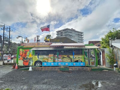 The image showcases a green, food-truck-like structure, emblazoned with the words "TOSTADURÍA 'La Molienda'", located in what appears to be Osorno, Chile, identifiable by the Chilean flag waving atop it. The food truck is adorned with images of various food items, including coffee beans, bread, and sandwiches. The side of the truck advertises a menu featuring items like "CAFÉ-TE", "CAFÉ DE C", "CHOCOLATE", and "LECHE CON MANI", along with "Mote con huesillo", a popular Chilean drink. The truck is elevated on a wooden platform, and a sign featuring the word "Savory" repeats along its base.
The surrounding environment consists of a gravel parking area, with visible signs of moisture, suggesting recent rain. In the background, a modern apartment building can be seen, providing an urban context to the scene. Utility poles and power lines are visible in the periphery. The presence of additional structures, including what appears to be a greenhouse or garden area to the right, suggests the food truck is situated in a mixed urban and semi-rural environment. The sky is partially cloudy. There is no evident people.