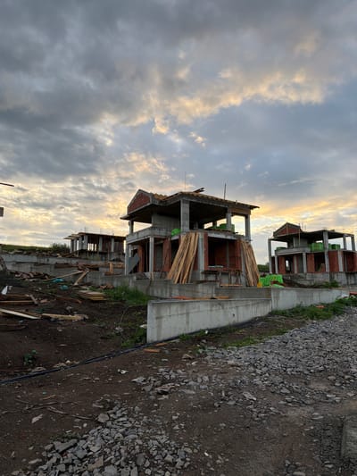 The image shows several unfinished buildings in Cherni Vrah, Bulgaria under a cloudy sky with patches of sunlight. The buildings are in the early stages of construction, with concrete frames and some brickwork visible. There is construction debris scattered around the site, including lumber and other materials. The ground is a mix of dirt and gravel. The image is taken from a low angle. There are no people visible in the image.