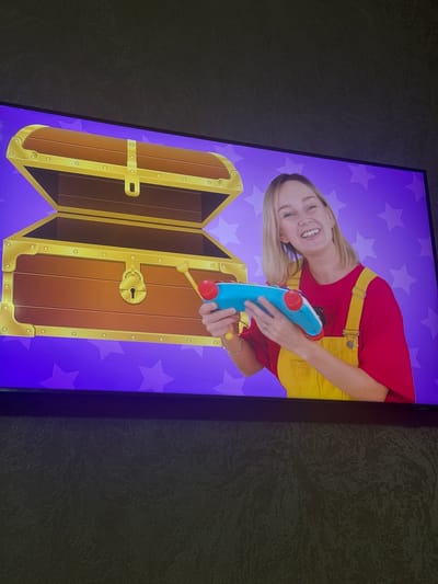 A blonde woman wearing yellow overalls over a red shirt smiles at the camera while holding a blue toy with red accents in front of an open, cartoonish treasure chest. The background is purple with white stars. The scene appears to be on a television screen, as indicated by the black frame and textured surface below. There is no discernible information to determine the location in Dushanbe, Tajikistan.