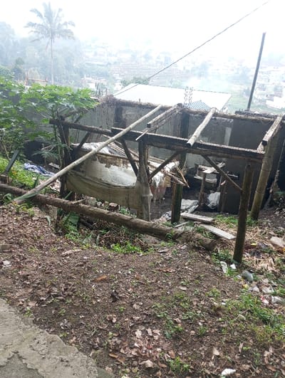 This image captures a dilapidated, partially constructed or ruined structure situated on a sloping, unkempt hillside in Gadog, Indonesia. The foreground consists of reddish-brown earth covered with fallen leaves, sparse green weeds, and scattered refuse including plastic bags and wrappers. A concrete edge is visible in the lower-left.
The central subject is an incomplete building featuring concrete block walls and an exposed framework of weathered wooden beams and poles, some appearing to be bamboo, indicating a lack of a roof. A white fabric or tarp is draped loosely within the structure, and various debris, including a white bucket, is visible inside. A long wooden pole extends diagonally from the structure towards the foreground.
In the mid-ground left, green foliage and a small tree are present. The background reveals a hazy, descending view of a populated area with numerous buildings, characterized by light-colored roofs, nestled within a valley. A tall palm tree stands prominently on the left hillside. Wisps of smoke are visible rising from a distant structure in the center background. Utility lines traverse the upper-right portion of the frame. No individuals are present, and no discernible actions or interactions are depicted. The scene is illuminated by ambient daylight under what appears to be an overcast or hazy sky.