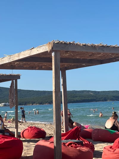 The image depicts a sunny day at a beach, likely in Primorsko, Bulgaria, featuring a wooden structure with a thatched roof, providing shade for beachgoers. Red beanbag chairs are scattered on the sandy beach, some occupied by people relaxing. The sea is visible in the background, with several individuals swimming and enjoying the water. A lush, green forested hill forms the backdrop of the scene, with a few white buildings visible on the left side, suggesting a proximity to a resort or town. The sky is a clear, bright blue. A towel hangs from the wooden structure, and a white tag with the number "3" is attached to one of the support posts.