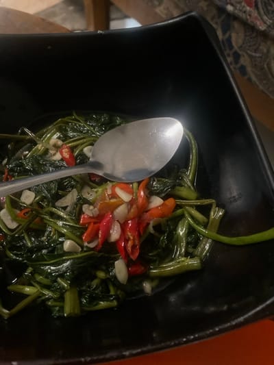 A black, square-shaped serving bowl contains a prepared dish of dark green, stir-fried water spinach (kangkung), visibly intermingled with numerous sliced red chili peppers and chopped white garlic cloves. A single silver-colored metal spoon lies horizontally across the top surface of the food. The background is blurred, revealing a light brown, possibly wooden, surface in the upper left, a patterned textile in the upper right, and a solid orange surface in the lower right, consistent with an indoor dining environment. This scene is located in Cimenyan, Indonesia.