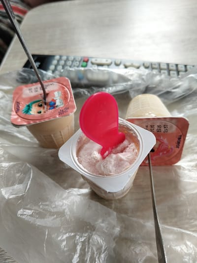 In this close-up, high-angle shot, three small, individual servings of a frozen dairy dessert, likely yogurt or ice cream, are arranged on a clear plastic sheet. The dessert in the foreground is pink and appears to be partially eaten, with a bright pink plastic spoon lodged in it. The lid of this container is white. To the left, another container, also partially eaten, has a pink lid featuring a cartoon character with green hair. A metal utensil is inserted into this container. To the right and slightly behind, a third, whole container of the dessert is visible, its lid a pale yellow color with visible text. The dessert itself appears creamy and light-colored. The background shows a blurred image of a remote control, suggesting an indoor setting. The overall impression is a casual moment of enjoying a frozen treat, possibly at home. The visible text on the lids includes "ТВОРОГ" (Tvorog - meaning quark or cottage cheese in Russian), and dates "07 05 26" and "15 06 26 A", indicating production or expiry information. The context of Shchastia, Ukraine, places this image in a specific geopolitical area, though no visual cues in the image itself directly confirm this location.
