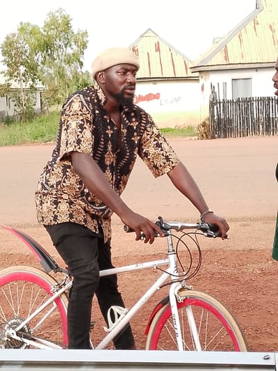 A man wearing a tan beanie and a patterned black and gold shirt is posing on a white bicycle with red rims. He has a beard and is looking to the right, with his hands on the handlebars. The man is outdoors on a dirt road. In the background, there are two buildings with light-colored walls and corrugated metal roofs, one of which has the word "Lodge" scrawled in red on its side. A fence made of vertical wooden poles is also visible. Green trees and grass are present on the left side of the image. The scene appears to be set in Agwala, Nigeria, during daylight.