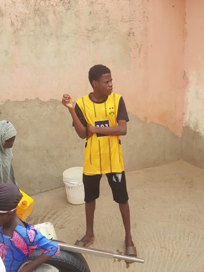 A young man wearing a yellow and black soccer jersey stands in the corner of a room or courtyard in Dutsinma B, Nigeria. The walls are painted in shades of pink and tan, and show signs of age and wear. The ground is a similar tan color. Near the man's feet are a white bucket and a tire leaning against a metal object, possibly a stand or tool. To the left of the tire, partially obscured, are figures lying on the ground; one person is covered in a blue and pink patterned fabric and another in a light-colored, patterned headscarf. A yellow container is also visible. The overall scene suggests a domestic setting or an informal gathering.