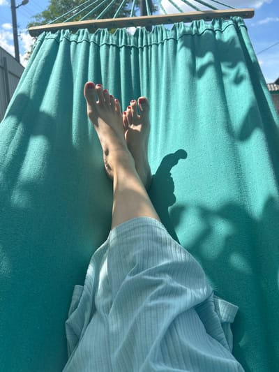The image shows a close-up view of a person's legs and feet lying in a teal-colored hammock. The person's feet are bare, with toenails painted red. They are wearing a light blue, ribbed garment that drapes over their legs. Sunlight casts shadows on the hammock, suggesting it is daytime, likely outdoors. The background is blurred but shows a glimpse of blue sky with white clouds, trees, and what appears to be a building or fence. The overall impression is one of relaxation and leisure. There is no visible text in the image. The context provided indicates the location is Vyshevychi, Ukraine.