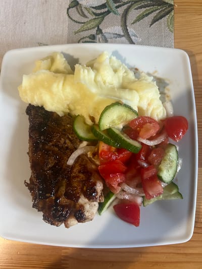 A white square plate holds a meal on a wooden table. The plate is positioned on a placemat with a subtle pattern of green leaves and branches. On the left side of the plate is a piece of dark, glazed meat, possibly chicken or pork, with visible char marks. To its right, a generous portion of mashed potatoes is piled high, appearing creamy and slightly lumpy. In the center and right of the plate is a fresh salad composed of chopped tomatoes, sliced cucumbers, and thinly sliced red onion. The salad is lightly seasoned with pepper. The meal is presented in a close-up, overhead shot, with natural light illuminating the food. The environment appears to be an indoor setting, possibly a restaurant or home dining area. The location in Zakopane, Poland is not discernible from the image.