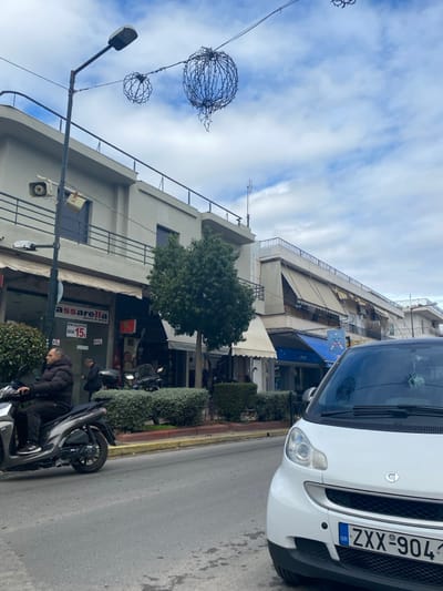 The image captures a daytime street scene in an urban area, consistent with Koridallos, Greece. The perspective is looking along a street lined with buildings and shops. The sky is overcast with visible clouds.
In the foreground, a white Smart car is partially visible on the right, parked alongside the curb. Its front license plate, facing the viewer, reads "GR ZXX-904." To the left of the car, a black scooter is parked, and a man wearing a dark puffer jacket is interacting with it, possibly getting on or off. The street itself is asphalt with a reddish-brown curb and green hedges separating the pavement from the road.
The midground features multi-story buildings housing various businesses. One prominent shop sign clearly displays "assarella" in red lettering, with "15€" and "DIA" visible below it, suggesting a price or offer. Another shop sign nearby has some Greek text. The buildings have multiple floors with balconies and awnings. Overhead, spherical wire decorations, possibly for festive lighting, are strung across the street, attached to poles, one of which also holds a street lamp. Trees and additional shrubbery are scattered along the pavement.
The background shows more residential or commercial buildings under the cloudy sky. The overall impression is a bustling urban street with a mix of everyday activity and civic decorations.