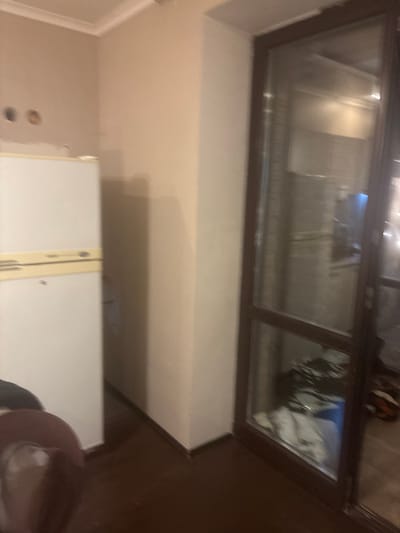 The image depicts an indoor scene, possibly a residential interior in the city of Orekhovka, Russia. On the left side of the frame, a white refrigerator is visible, with a yellow strip across its upper section. Above the refrigerator, two circular openings are present on a light-colored wall. To the right, a large sliding glass door with a dark brown frame dominates the scene. The glass reflects the interior, showing what appears to be kitchen or living area furnishings, including a counter and some cabinets, with a blue object and some white fabric visible near the bottom of the reflection. The floor is dark brown. In the foreground, partially visible on the lower left, is a section of a dark-colored object, possibly furniture or clothing. The lighting suggests an indoor environment, with light sources reflecting off the surfaces.