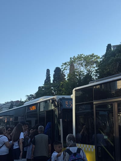 In Şişli, Turkey, on a clear day, a group of people are gathered around a bus. The bus displays a digital destination board with Cyrillic characters. Some individuals are standing in front of the bus, while others are entering or exiting. The scene appears to be a bus stop, with several passengers waiting for transportation. The surrounding environment includes trees and buildings.