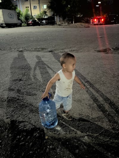 In the city of Şamaldı-Say, Kyrgyzstan, a young child, appearing to be a toddler, is depicted walking on a gravelly surface at night. The child, wearing a light-colored tank top and denim shorts, is carrying a large, blue, plastic water bottle with a blue cap. The child's right hand grips the bottle's neck. The child is looking towards the right side of the frame, with their body angled slightly away from the viewer. The ground is uneven, with visible pebbles and tire tracks. Long shadows are cast on the ground, suggesting a light source from behind and to the left of the child. In the background, the blurry shapes of buildings and vehicles are visible. The lighting in the scene is dim, with artificial light sources creating red hues and reflections.