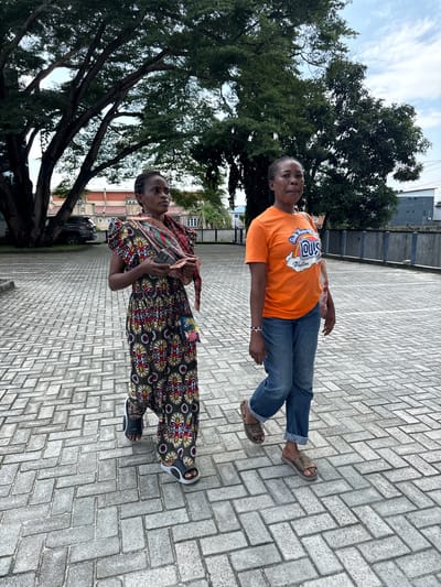 The image shows two women standing on a paved walkway. The woman in the foreground is wearing a colorful, patterned dress and a scarf around her neck. She is holding a phone in her right hand and looking to her left. The woman in the background is wearing an orange t-shirt with a graphic design and blue jeans. She is standing with her arms at her sides and looking to her left.
The setting appears to be an outdoor area with trees and buildings in the background. The ground is paved with interlocking brick pavers. The sky is visible, suggesting it is daytime. The scene is likely taking place in Epe, Nigeria, as indicated by the location context. There is no clear indication of a specific event or activity beyond the two women standing and posing for the photo.
Notable details include the clothing of the women, the patterned dress and the graphic t-shirt, as well as the brick-paved walkway. The text on the orange t-shirt is partially visible and appears to be a logo with colorful lettering. The overall mood seems casual and relaxed.
