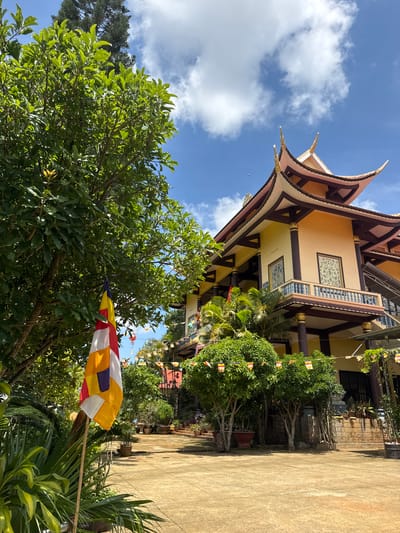 The image captures a vibrant scene in Lạc Dương, Vietnam. A temple, likely a Buddhist temple, is partially visible in the background with a distinctive multi-tiered roof and beige walls accented with dark brown beams and decorative windows. Several colorful Buddhist flags are strung around the temple grounds. In the foreground, lush greenery dominates, including a large tree with thick foliage and various potted plants. A striped Buddhist flag is prominently displayed, its colors of red, white, yellow, and orange catching the sunlight. The sky is a mix of blue and white clouds.