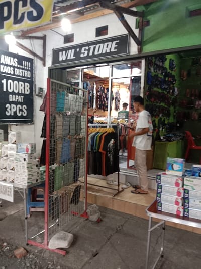 The image shows a storefront with the sign "WIL'STORE" in Kaliwungu, Indonesia (latitude: -6.962, longitude: 110.252). The storefront is open, and a man in a white shirt and khaki pants is standing in the doorway, looking at a rack of clothing inside. The rack is filled with t-shirts. There are other items of clothing visible on the walls inside the store, which has a tiled floor. There is a red and black metal rack outside the storefront, displaying an assortment of face masks for sale. A folding table with a stack of boxes containing medical supplies is positioned to the right of the rack. There is a red plastic chair and a small blue chair positioned in front of the table. There are two large rocks on the sidewalk in front of the store. A sign with the text "AWAS....!!! KAOS DISTRO 100RB DAPAT 3PCS" is visible on the left-hand side of the storefront. The sidewalk is made of concrete, and there are some loose bricks and stones in the foreground. The building has a white exterior and a green exterior wall on the right side of the building.
