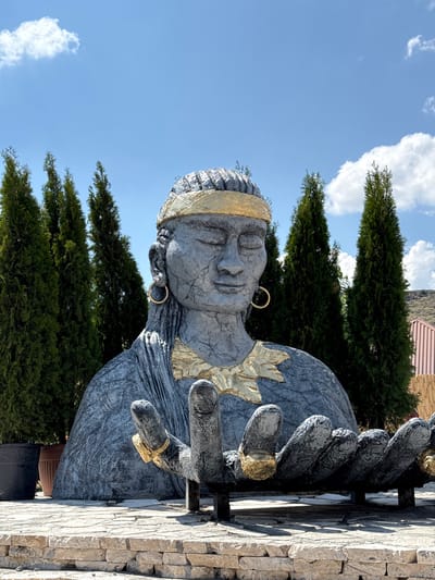 This is a frontal, eye-level, medium shot of a large, gray, stone statue of a person with closed eyes, wearing a gold headband and gold jewelry. The statue is depicted from the chest up, with its large hands extended forward, also adorned with gold rings. The statue is situated outdoors, in front of a line of tall, dark green cypress trees. Behind the trees, a portion of a reddish-brown building with a thatched roof is visible on the right side of the image, along with a hillside. The sky above is blue with scattered white clouds. The statue is placed on a stone-paved area. This is the Armenian Mother sculpture, located in Ptghni, Armenia.