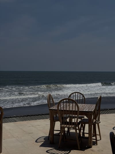 The image displays an outdoor patio scene in Belalang, Indonesia, facing a dark sand beach and ocean under a dark, uniform sky. In the foreground, a light-toned tiled patio floor supports a rectangular wooden table with a patterned surface and four matching wooden Windsor-style chairs, arranged in a dining configuration. Shadows from the table and chairs are cast onto the patio, suggesting direct sunlight. A black mesh net structure is visible along the edge of the patio, bordering the dark sand of the beach. The midground shows ocean waves breaking with white foam near the shore. The background is composed of the open ocean meeting a largely overcast, dark grey sky. No individuals are present in the scene.