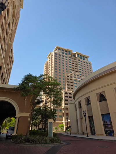 This image captures a street view in the Pearl-Qatar, a mixed-use development in Doha, Qatar. The scene is dominated by modern architecture, characterized by beige-colored buildings with arched windows and balconies. A tall, multi-story residential tower stands prominently in the background, its facade featuring numerous windows. In the foreground, a curved, low-rise building with arched entrances and ornate detailing lines the street. A tree with green foliage provides a natural element amidst the urban landscape. The street itself is paved with reddish-brown bricks, and lampposts line the sidewalk. A portion of an arched entryway is visible on the left, revealing a glimpse of a vehicle and more greenery beyond. The sky is clear and blue, indicating a sunny day.