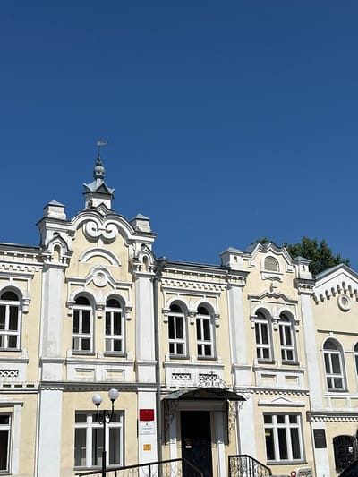 The image captures the facade of a pale yellow, two-story building with white decorative elements, set against a clear, bright blue sky. The building exhibits an ornate architectural style, with numerous arched windows on both levels. The upper facade features elaborate cornices, decorative moldings, and a prominent central gable with a weather vane topped with the numerals "1883". A portion of green foliage from a tree is visible on the right side of the frame.
The building is located in Biysk, Russia, and appears to be a public or institutional building. A sign with Russian text and a red rectangular plaque are mounted on the wall near the entrance on the ground floor. A black door with a decorative wrought-iron awning is visible, as are a pair of white globe-shaped streetlights. A black metal railing with an ornate design lines the stairs leading to the entrance. In the lower right corner, a partial view of a vehicle and a segment of a red and white sign with the word "moxim" are visible. The scene is static, depicting the exterior of the building on a sunny day.