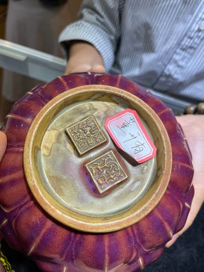 In the Futian District of China, a person's hands are holding a purple, fluted ceramic bowl. Inside the bowl, two small, square bronze seals rest on a lighter-colored ceramic base. The seals appear to have intricate carvings on their surfaces. A small, red-and-white tag with Chinese characters written in black ink is placed on top of one of the seals. The person is wearing a light-colored, striped shirt. The lighting suggests an indoor setting, possibly a shop or a private collection display. The overall scene conveys a sense of appraisal or examination of these artifacts. The text on the tag is "龚开门".