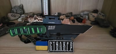 The image shows a tactical knife with a black blade and a green and black patterned handle. A small red dot sight is mounted on the blade. The knife is resting on a black sheath. In front of the knife, there are two patches: one is a small Ukrainian flag, and the other is a patch with the letters "G.I.A.N.K.E.R." The background consists of various rocks and a thermometer. The scene appears to be set on a wooden surface, possibly a shelf. There is no indication of the city of Pabianice, Poland.