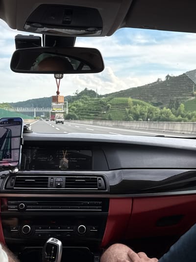 The image is taken from the interior of a car driving on a highway. The dashboard features a navigation system displaying a route, a car stereo, and climate controls. A phone is mounted on the dashboard. A rearview mirror is visible, reflecting the sky and a person's head. A car air freshener hangs from the mirror. Outside the car, a highway stretches ahead, flanked by green, terraced hills. Several vehicles are visible on the road. The sky is partly cloudy. The location is identified as Fuyang, China.