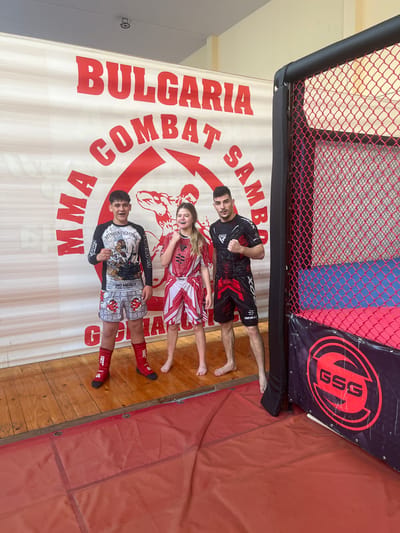 This media file shows three individuals, two males and one female, posing in front of a banner that reads "BULGARIA COMBAT SAMBO MMA". The setting appears to be an indoor combat sports training facility, possibly a gym or ring area, given the presence of a chain-link fence in the background and a red mat on the floor. The banner itself is large and white with red lettering, featuring graphics of muscular figures. A logo with the letters "GSG" in a red circular emblem is also visible on a black banner above the main one.
The individuals are dressed in athletic wear suitable for combat sports. The male on the left is wearing black and red shorts and a matching short-sleeved rash guard. The female in the middle is wearing a red and white patterned outfit, possibly a martial arts uniform. The male on the right is wearing black and grey shorts with a long-sleeved shirt and red shin guards. All three individuals are smiling and appear to be in good spirits, suggesting a posed photograph rather than an active competition. The lighting indicates it is daytime. The location context provided is Gorna Oryahovitsa, Bulgaria.