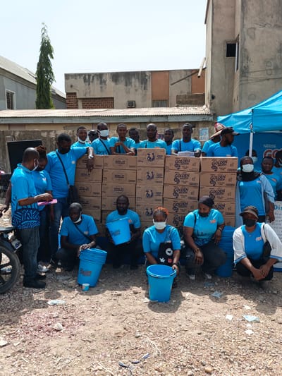 The image shows a group of approximately 20 people in Kawaji, Nigeria, likely involved in a promotional or distribution event. Most of them are wearing light blue t-shirts with the "Golden Morn" logo, with some also wearing face masks. They are gathered behind and beside a stack of brown cardboard boxes labeled "Bowl of Goodness" and "Golden Morn." In front of the boxes, several people are either kneeling or crouching next to light blue buckets with the "Golden Morn" logo. The ground is a mix of dirt, gravel, and scattered debris. Behind the group, there is a partial view of a blue promotional tent with a "Nestle" logo, sections of buildings, and a motorcycle parked to the left. The overall setting appears to be an outdoor space in a residential or commercial area. The sky is bright and mostly clear.