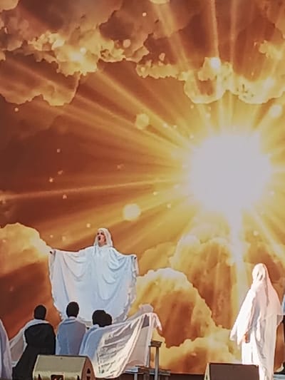 The image depicts a theatrical performance, possibly with religious or allegorical themes, set against a dramatic backdrop. The scene is dominated by a large, stylized sunburst emanating from the upper right quadrant, with radiating beams of light casting a warm, golden glow across clouds. These clouds appear to be part of a painted backdrop, rendered in shades of orange and yellow, creating a celestial or divine atmosphere.

In the foreground, several individuals are present. The central figure is a person dressed in a flowing white robe, with arms outstretched and mouth open as if singing or speaking. Their blonde hair is visible, and the overall presentation suggests a significant role, perhaps angelic or divine. To the left and behind this central figure, three individuals are seen from behind, dressed in dark and light blue attire. They appear to be seated or kneeling, facing the main performer, their posture suggesting an audience or participants in the narrative. On the far right, another person in a white hooded robe stands, facing away from the camera, adding to the ensemble.

The stage itself is partially visible, with a dark structure in the lower left and a white structure or prop to the right. The lighting is intense, particularly from the sunburst, creating a bright, almost ethereal effect. The overall mood is one of awe, reverence, or intense emotion, amplified by the dramatic lighting and the stylized celestial backdrop. There is no visible text in the image. The context provided places this event in Yaroslavl, Russia.