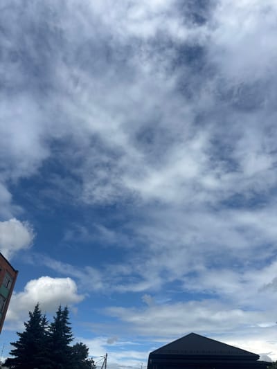 The media file is a low-angle, vertical shot of a cloudy blue sky. Wispy white clouds stretch across the frame, with darker patches indicating denser cloud formations. In the upper right corner, the dark, angular roof of a building is visible, suggesting an urban or suburban setting. Towards the bottom right, the dark green, spiky branches of a coniferous tree are prominent. A section of a brick building with green window frames can be seen in the lower right quadrant. The lighting suggests it is daytime, with bright sky and shadows. The overall mood is natural and perhaps a bit overcast. No text is visible in the image. The location context provided is Капыльскі сельскі Савет, Belarus, which aligns with the presence of typical European architecture and vegetation.