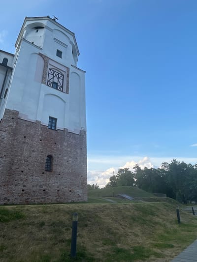 The image depicts a stone and white tower with a blue sky backdrop. The tower has a brick lower section and a white upper section. There are several windows visible on the tower. In front of the tower, there is a grassy area with some landscaping and trees in the background. The scene appears to be outdoors, possibly in a historical or park setting. The overall impression is one of a sunny day with a clear sky and some clouds.