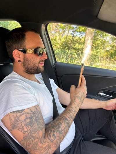 A man is sitting in the passenger seat of a car, looking at his phone. He has a beard, sunglasses, and a white t-shirt. He has tattoos on both arms. The car is moving, as can be seen by the trees outside the window. The location is not possible to determine without additional information.