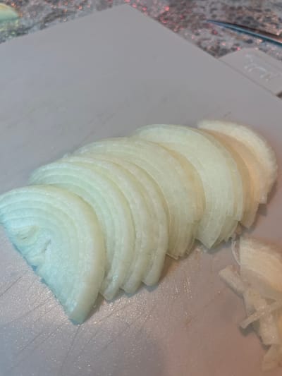 The image shows a pile of thinly sliced white onions on a white cutting board. The onions are stacked in a slight curve. Some loose onion pieces are scattered to the right of the stack. A small portion of a counter, possibly granite or a similar material, and what appears to be a kitchen utensil are visible in the upper left corner. The image does not provide information about the location being in Olginka, Russia.