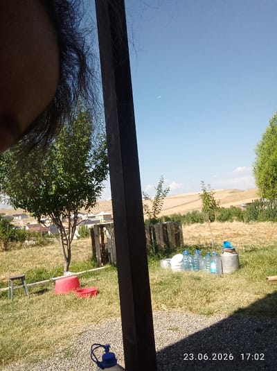 The image depicts a scene outdoors, likely in a rural or semi-rural setting. In the foreground, a dark vertical structure partially obstructs the view. To the left, a portion of a person's face and dark hair is visible, with the person appearing to be looking towards the right or slightly down.
The midground shows a grassy area with several objects. A small tree with green foliage stands in the center. To its left, there is a small green stool or step. Two red containers, one larger and one smaller, are present on the grass near the tree. Further to the right, a row of plastic water bottles with blue caps is lined up, along with a metal canister and a white bucket. Behind these items, a rustic wooden fence or structure is visible.
The background consists of dry, yellowish grass and some scattered green bushes. In the distance, there are rolling hills with patches of dry vegetation and a few buildings with light-colored roofs, suggesting a residential area. The sky is a clear, bright blue with a few faint white clouds. A tiny speck, possibly the moon, is visible in the upper left portion of the sky. The date "23.06.2026" and time "17:02" are superimposed on the bottom right of the image. Based on the provided information, this scene is located in the city of Mady, Kyrgyzstan.