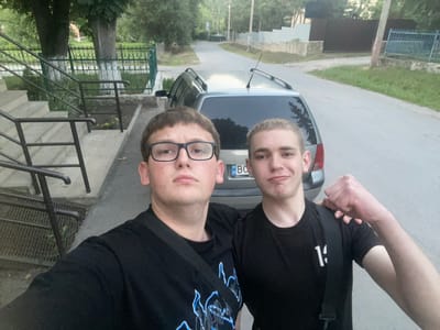 Two young men are posing for a selfie outdoors, with a silver station wagon parked behind them on a paved road. The man on the left is wearing glasses and a black t-shirt with a graphic design. The man on the right is wearing a black t-shirt with the number "14" on it and has his arm raised, showing a clenched fist. They are standing on a sidewalk next to a set of concrete stairs and a metal railing. In the background, there are trees, a green metal fence, and some buildings. The license plate on the car is partially visible and appears to be from Ukraine. The scene suggests a casual encounter in a residential or semi-rural area. The weather appears overcast, and the lighting indicates it is either early morning or late evening. The location is specified as Tovstoluh, Ukraine.