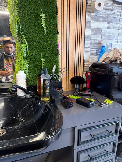 This image, oriented sideways, depicts the interior of what appears to be a modern barbershop or hair salon in Dragor, Bulgaria. The primary focus is a countertop and sink area filled with barber tools and products.
On a gray countertop, various items are arranged. There's a large black device, possibly a sterilizer or charging station, with "BOOST" visible on its side. Several red hair clippers are present, one mounted in a charging dock, another smaller one near a black brush, with the word "INSTINCT" visible on it. Yellow and green combs, a black round brush, and multiple pairs of scissors are also neatly laid out. A yellow keyring with a tag hangs from an upper cabinet.
To the left, a vertical wooden slatted panel serves as a wall, and a faux grass wall adorned with artificial green foliage provides a contrasting backdrop. Black and purple hair clips are placed on the grass wall.
A black sink with white marbling occupies the lower right portion of the image. On the edge of the sink, two brown bottles with pumps are visible, both bearing the label "RED ONE" and containing different liquid colors (one golden, one darker brown). A white bottle with a red label is also next to the sink's faucet.
Reflected in the mirror above the sink is a young man, likely the barber or a client, his face partially visible and his hands near his head, implying an ongoing or recently completed haircut. He is wearing a barber's cape.
In the upper background, a light-colored, patterned floor (resembling wood planks) is seen, along with a roll of paper towels and a blue spray bottle. Above the countertop, a gray cabinet with two framed doors and ornate black handles is visible. The overall scene suggests a functional and stylish barbershop environment.