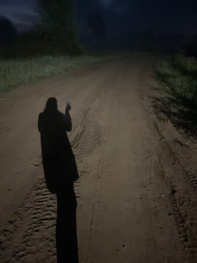 An unpaved road stretches into the darkness, lined by tall grass on either side. The shadow of a person, facing away from the camera and wearing a long coat, is cast on the road. The shadow is making a peace sign with its right hand. The overall scene is dark, suggesting it is nighttime. The specific location in the city of Уральское, Russia is not discernible from the image alone.