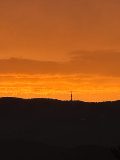An image of the sunset over Strazha, Bulgaria, depicts a striking sky filled with shades of orange and yellow. The sky is dominated by layers of textured clouds, some appearing wispy and others more solid, all illuminated by the setting sun. In the distance, silhouetted against the vibrant sky, is a prominent radio tower situated on a dark, forested hilltop. The foreground is a dense silhouette of trees and rolling hills, obscuring any details of the landscape below. The overall impression is one of natural beauty and tranquility as day transitions into night.
