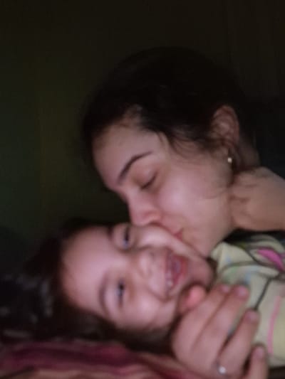 An out-of-focus, grainy image shows two individuals, a child and an adult female. The adult female, positioned on the left, has dark hair, and her eyes are closed. Her skin tone is light. To her right is a child smiling with visible teeth. The background is a dark green. It is not possible to identify any specific location in Byala Slatina, Bulgaria from this image.