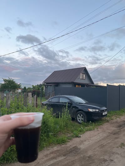 A hand holds a clear plastic cup containing a dark liquid and white foam. In the background, a gray, two-story house with a brown roof is visible. A dark-colored sedan is parked in front of the house. The scene is set in a rural area with a dirt road, overgrown grass, and a wooden fence. Overhead, power lines crisscross the sky, which is filled with clouds. The license plate of the car reads "Y798MA 56". The location is identified as Klobyushnevo, Russia.