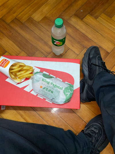 The image shows a person wearing black sneakers and black pants standing on a wooden floor in Llavallol, Argentina. A bottle of "Up" soda lies on the floor. Under the person's shoe is a red Nike shoebox, on top of which are a Burger King King Vegetal burger and a container of Burger King french fries.