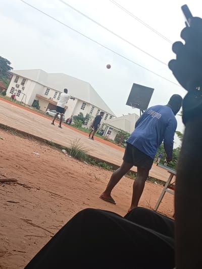 This media file captures an outdoor scene, likely on a basketball court, with several individuals engaged in an activity. The foreground is dominated by the legs and lower torso of a person sitting and looking out at the scene, with a black fabric covering their legs.
In the midground, a group of people are present on a reddish-brown court. One person, wearing a white t-shirt and dark shorts, is in motion, possibly throwing or receiving a ball, which is visible in the air above and slightly to the left of the court. Another person, wearing a blue long-sleeved shirt with white text on the back and dark shorts, is also on the court, facing away from the camera. A third person, in a dark t-shirt and shorts, is further down the court. In the background, there's a large, modern building with a white roof and multiple windows, possibly an institutional building or school. A white car is parked near the building. The court appears to have some infrastructure like a basketball hoop and backboard visible.
The time of day appears to be daytime, possibly overcast, given the bright but diffused light. The weather seems temperate. The ground is a mixture of reddish earth and some sparse vegetation. The scene suggests a casual sporting event or practice.
The text on the back of the blue shirt is partially visible and appears to be in English. While illegible in detail, it seems to be a statement or a slogan. The overall mood of the image is relaxed and active.