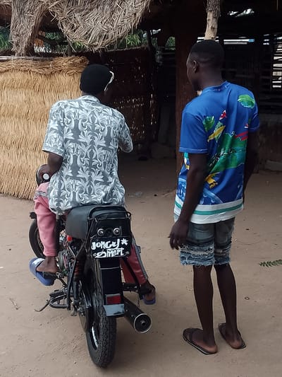 A low-angle shot depicts two dark-skinned African men in Biliri, Nigeria. One man sits on a black motorcycle, facing away from the viewer, wearing a gray patterned shirt and pink pants. A child wearing similar pants and a pink shirt sits in front of the man. The motorcycle's license plate reads "Joncy Smart" with a star graphic. To the right stands another man in a blue shirt with a landscape print and distressed denim shorts, facing towards the motorcyclist. Both men wear flip-flops. The scene is set outdoors on a dirt ground. Behind the men are thatched roof structures. The atmosphere seems casual.