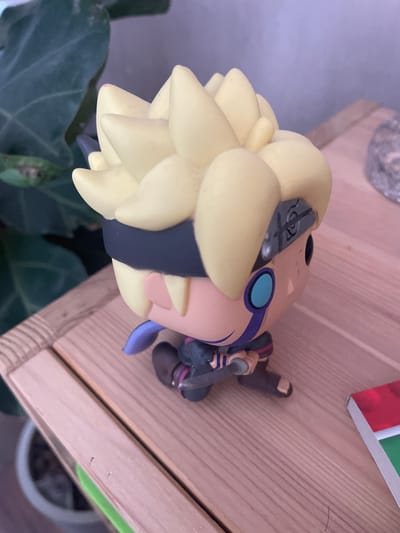 This is a close-up, horizontal, eye-level shot of a Funko Pop figurine of Boruto Uzumaki, a character from the anime and manga series *Boruto: Naruto Next Generations*. The figurine is primarily yellow with blue and purple accents, depicting Boruto with his characteristic spiky hair and a headband. He is shown in a dynamic pose, possibly mid-action, with one hand near his face and the other holding a weapon. The figurine appears to be made of vinyl.
The figurine is placed on a wooden surface, likely a table or shelf, with visible wood grain. To the left, a large, dark green leaf of a houseplant is partially visible, suggesting an indoor setting. In the background, out of focus, a light purple surface can be seen, possibly a wall. At the top right, a portion of what appears to be a book or box with red and green stripes is visible.
The lighting suggests an indoor environment, possibly with natural light. There is no indication of the time of day or weather conditions. The scene is static, with the figurine posing as an object rather than engaging in an activity. No text is clearly visible on the figurine or its immediate surroundings, other than the potential faint markings on the headband. The overall impression is that of a collector's display.