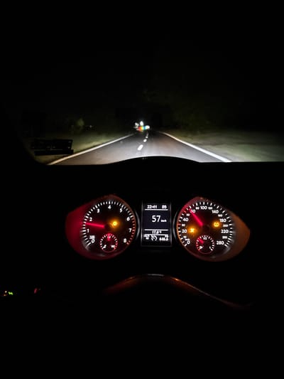 The image captures a night scene from inside a car, showing the illuminated dashboard and the road ahead. The perspective suggests the driver's view, with the dashboard prominent in the foreground and the road stretching into the darkness beyond the windshield.
On the dashboard, two primary analog gauges are visible. The speedometer, on the left, indicates a speed of 57 km/h, with a maximum reading of 280 km/h. A yellow warning light is illuminated within this gauge. To its right, the tachometer shows the engine RPMs around 1500, with a scale extending up to 8 x 1000 RPM. Another yellow warning light is active here.
Below these gauges, a digital display provides additional information: the current time is 22:41 (10:41 PM), the car is in 5th gear ("05"), the speed is reiterated as 57 km/h, the outside temperature is 17.0 °C, and a trip meter reads 646.6 km. A segment of the steering wheel is visible on the right side of the dashboard.
The view through the windshield reveals a dark night road, illuminated by the car's headlights. White lines clearly mark the two lanes of the road, and grassy verges are visible on either side. In the far distance on the left side of the road, faint, multi-colored lights suggest the presence of another object or person, possibly a bicycle with reflective elements or a small vehicle. This scene takes place in Dolna banya, Bulgaria.