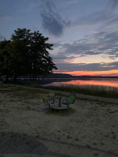 This image depicts a waterfront playground scene at dusk. In the foreground, a circular merry-go-round with four colorful seats (two yellow, two green) is situated on a sandy surface with sparse grass. To the left, a large tree casts a shadow. In the background, a calm lake reflects the vibrant colors of the sunset. The sky above is a gradient of deep blues and grays, transitioning into warm oranges and pinks near the horizon. Hills line the opposite shore of the lake, with scattered lights indicating a distant settlement. The overall impression is one of a serene, post-sunset ambiance in an outdoor recreational area. The location is the waterfront park in Berezhany, Ukraine.