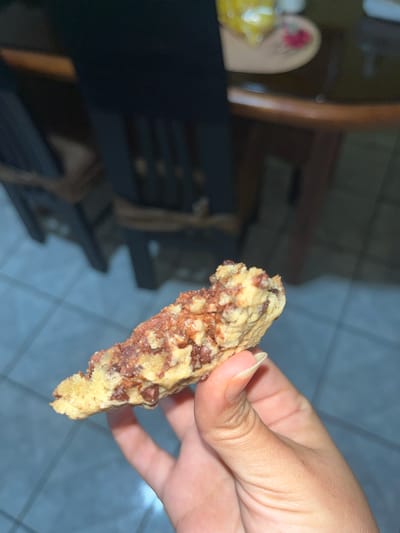 A close-up shot shows a hand holding a broken piece of a chocolate chip cookie. The cookie appears to be homemade with visible chocolate chips and a slightly irregular texture. The background is blurred, but hints at a domestic setting with furniture, possibly a dining table and chairs. The lighting suggests an indoor environment, likely during the day due to the presence of natural light. There is no indication of the specific location within San Luis, Peru.