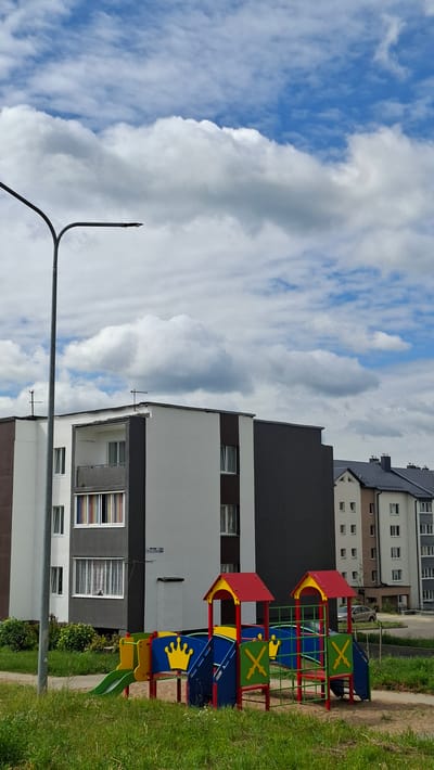 The image captures a brightly colored playground in Smalyavichy, Belarus, juxtaposed with modern apartment buildings under a cloudy sky. The playground features multiple slides, climbing structures, and platforms in red, yellow, blue, and green. A prominent yellow crown design is visible on one of the blue play areas. The apartment buildings are contemporary in design, with varying shades of grey, white, and brown facades. Some buildings have balconies, and the windows are visible. A tall street lamp stands in the foreground, its metal pole extending upwards. Lush green grass borders the playground and the sidewalk. The sky is a mix of bright blue patches and large, billowing white clouds, suggesting it is daytime, possibly with intermittent sunshine. The overall scene conveys a sense of urban residential life with amenities for children. No people are visible in the frame. There is a small sticker with text on the building's facade, but it is illegible.