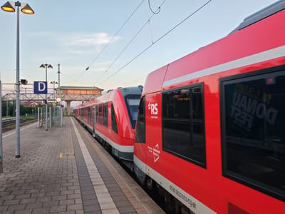The image depicts a train station in Memmingen, Germany, at dusk. Two red regional trains are stopped at a platform. The train on the right, closer to the viewer, has its doors closed. The train on the left is partially visible behind the first train. The platform is paved with gray and beige interlocking bricks. There are streetlights with orange lamps, a sign with the letter "D" in a blue square, and overhead electrical wires for the trains. In the background, a pedestrian bridge crosses the tracks, and buildings with warm lighting can be seen. A person is sitting on a bench at a shelter on the platform. The sky is a pale blue with wispy clouds. Reflections are visible in the windows of the train. The overall impression is of a quiet moment at a train station as evening approaches.